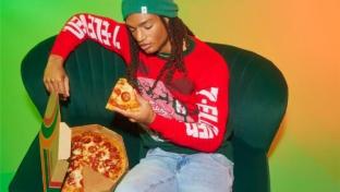 Man in a 7-Eleven sweater and beanie eating a 7-Eleven pizza