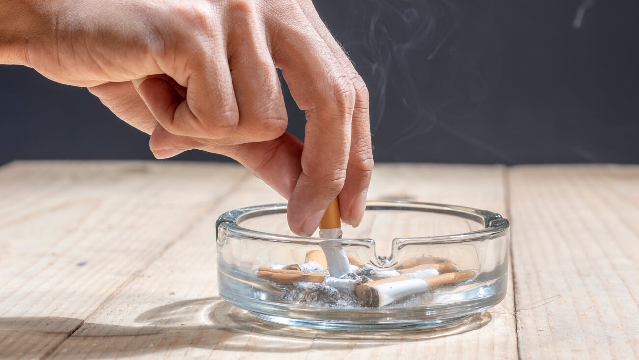 Hand stubbed out cigarette in a transparent ashtray on wooden table; Shutterstock ID 1440960986