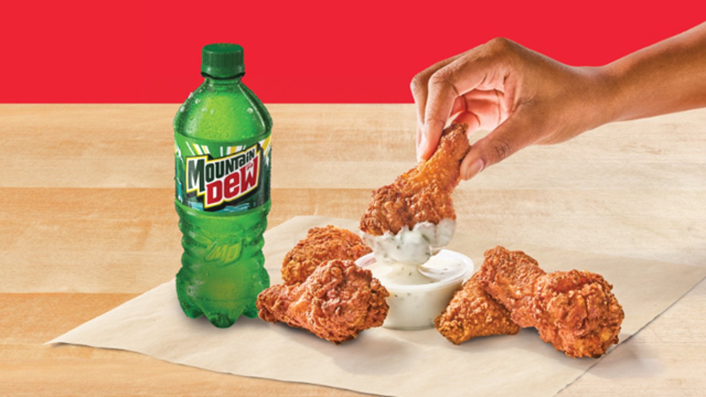 A person dipping chicken wings in ranch dressing with a Mtn Dew soda
